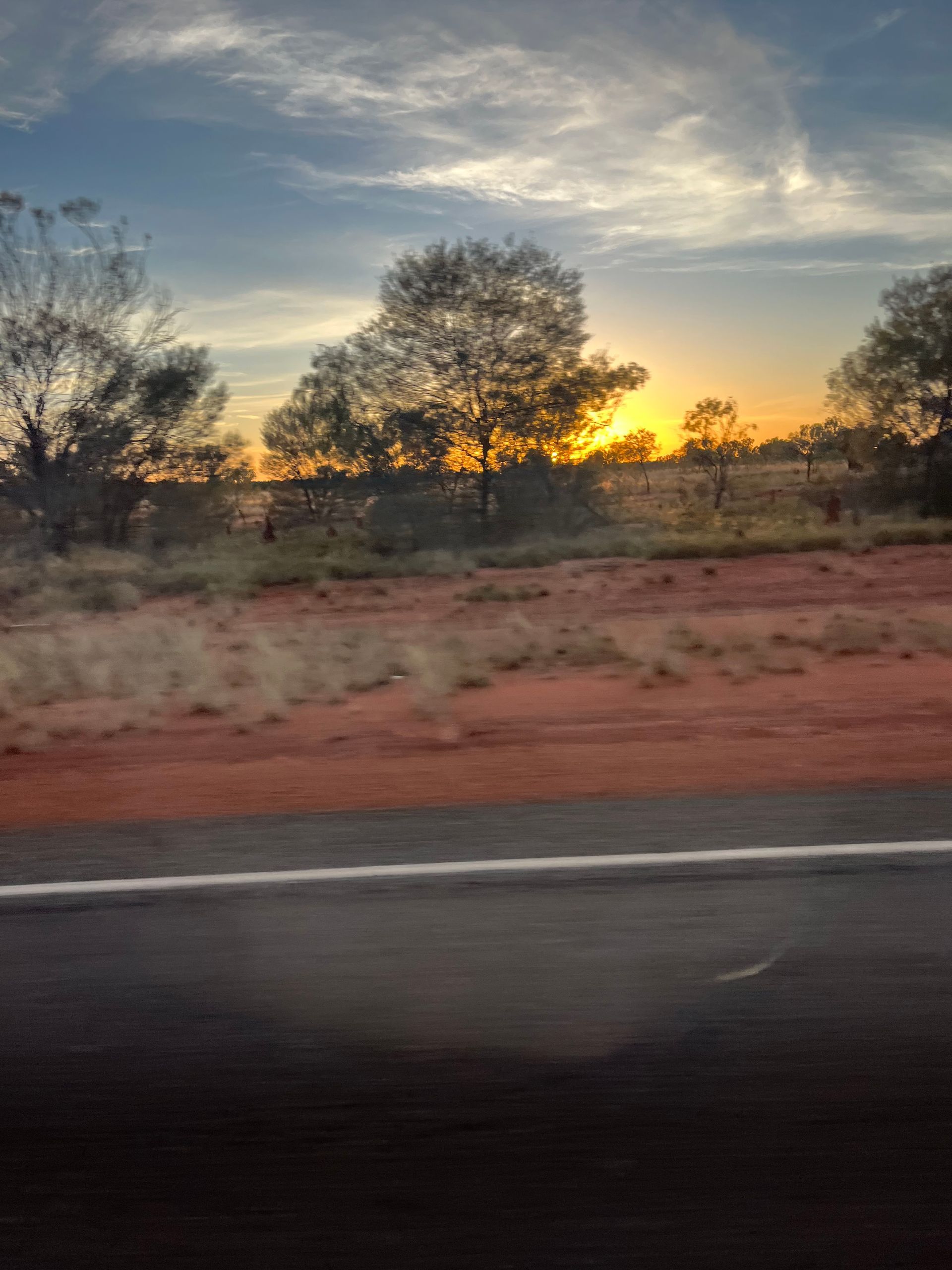 Sunset over a rural roadside with silhouetted trees and red dirt under a cloudy sky