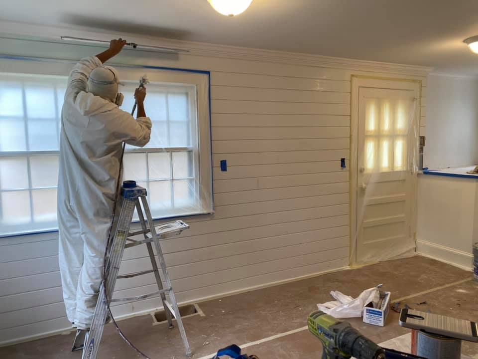 Person in protective suit sprays white paint on a shiplap wall inside a room, masking on windows and doors.