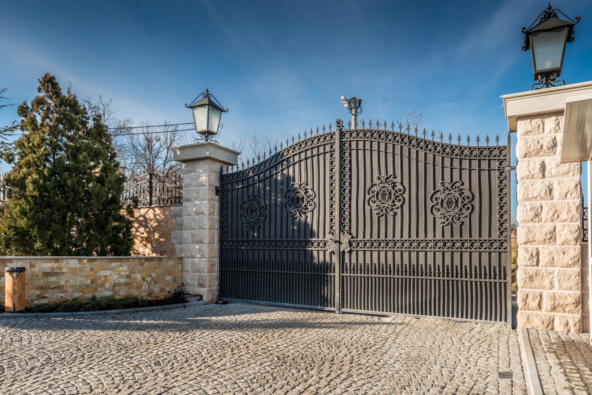 Ornate black wrought-iron gate between stone pillars under a blue sky, with gravel foreground.