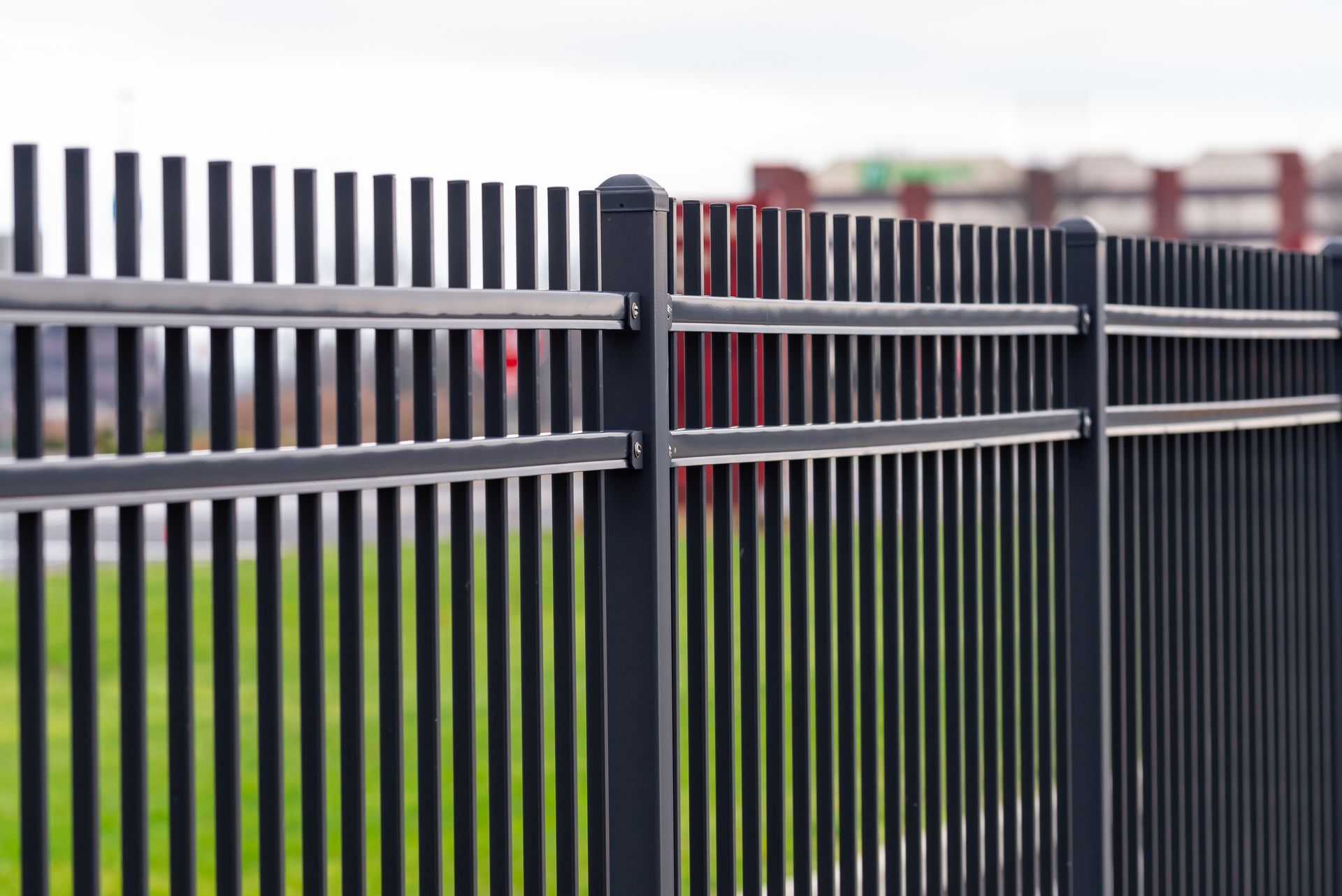 Black metal fence with vertical bars running along a grassy area with buildings blurred in backgroun
