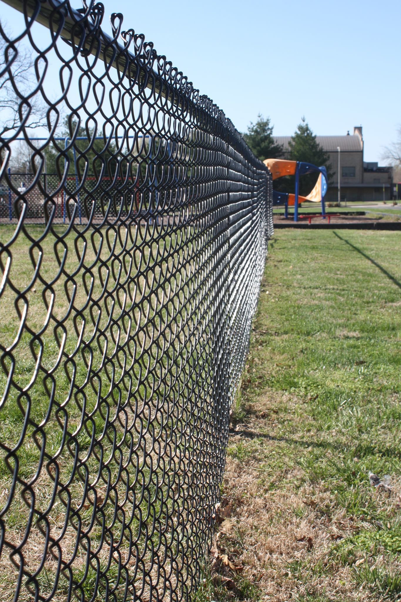 Chain-link fence beside a grassy field on a sunny day, with playground equipment and trees in the distance.