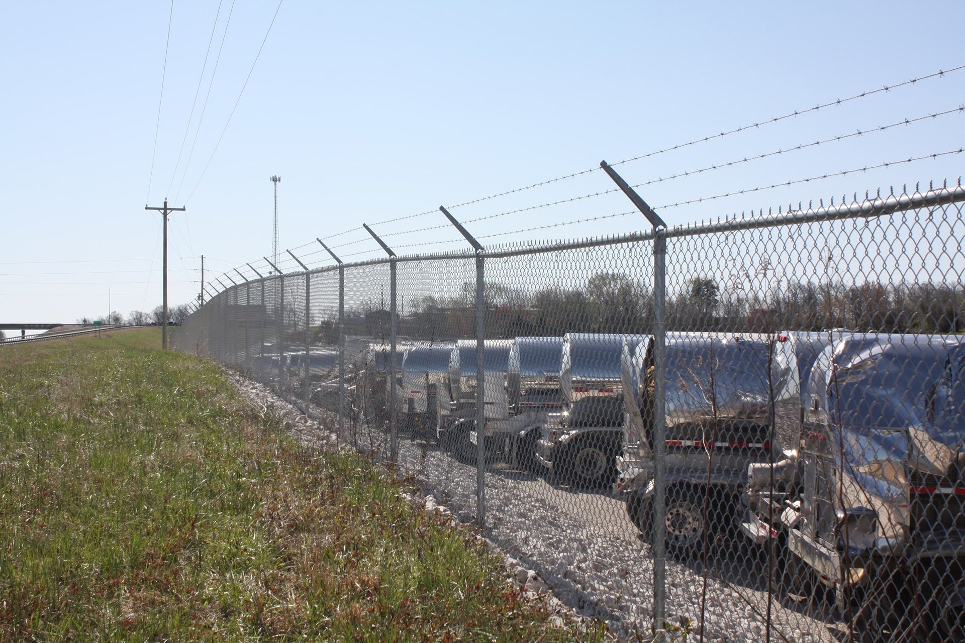 Rows of parked vehicles behind a chain-link fence along a grassy roadside under a clear sky