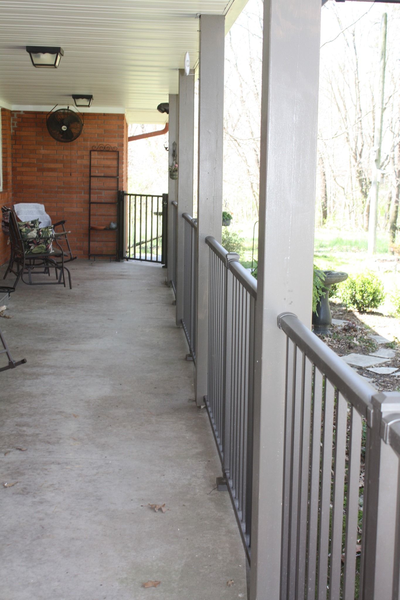 Covered porch walkway with gray railing, brick wall, and outdoor chairs beside a yard with trees