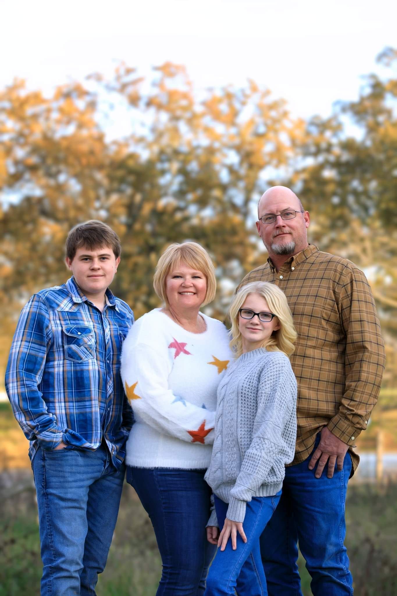 Family of four standing outdoors in autumn, with trees and soft golden leaves in the background.