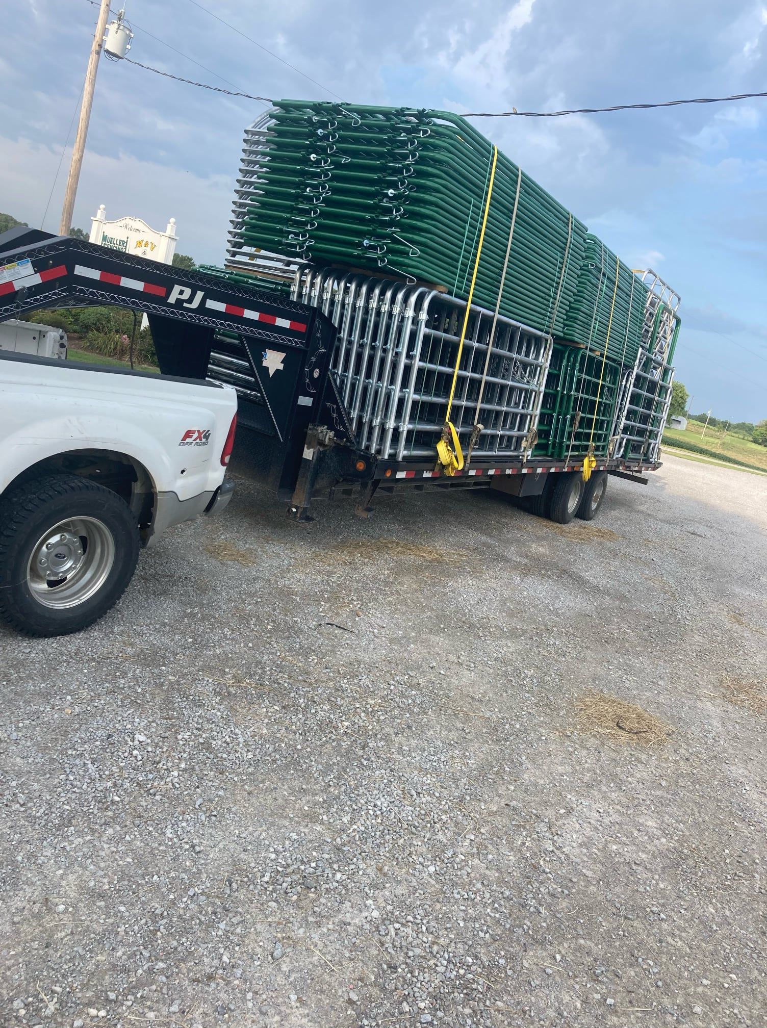 White pickup towing a flatbed trailer loaded with stacked green plastic pallets on a gravel lot under a blue sky