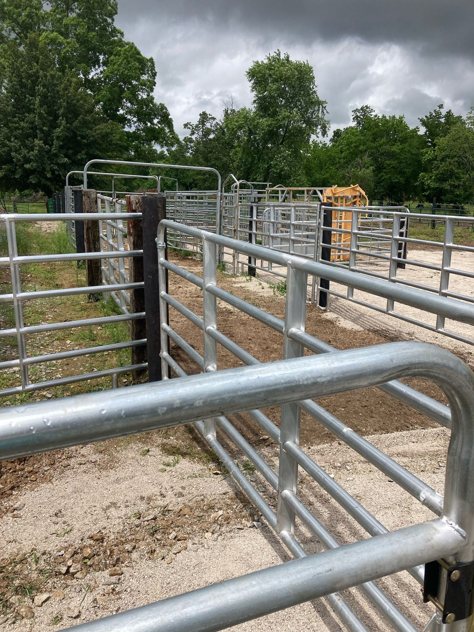 Metal livestock gate in a fenced corral under cloudy sky, with trees and dirt ground.
