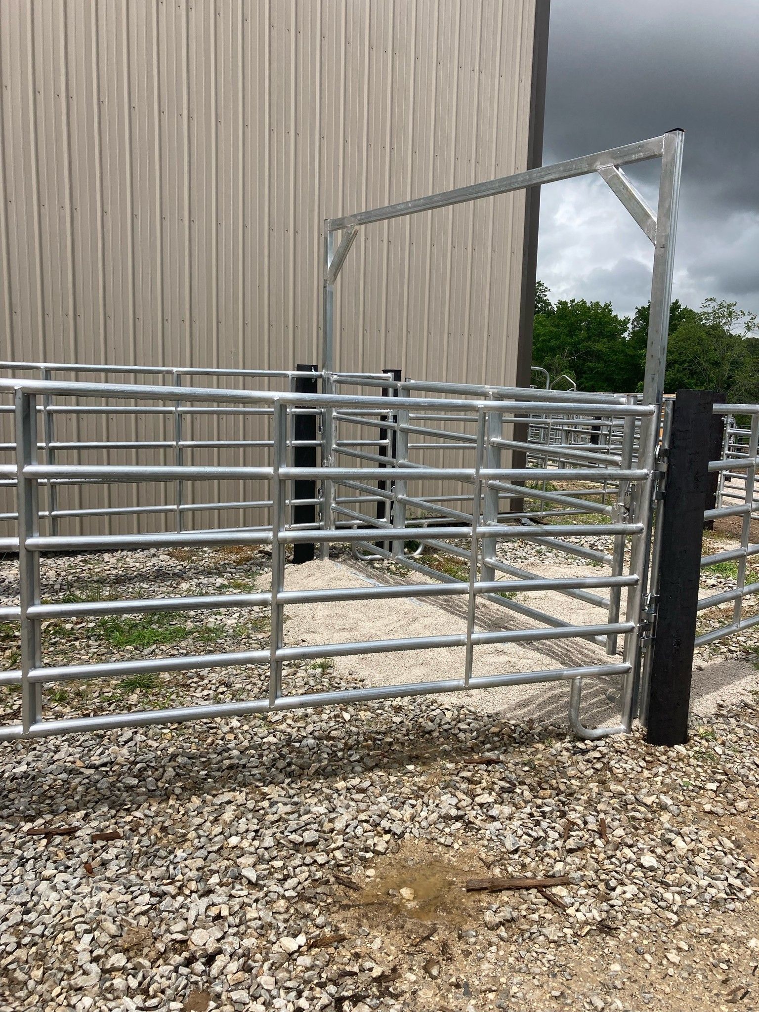 Metal livestock gate beside a beige barn, with gravel ground and trees in the background