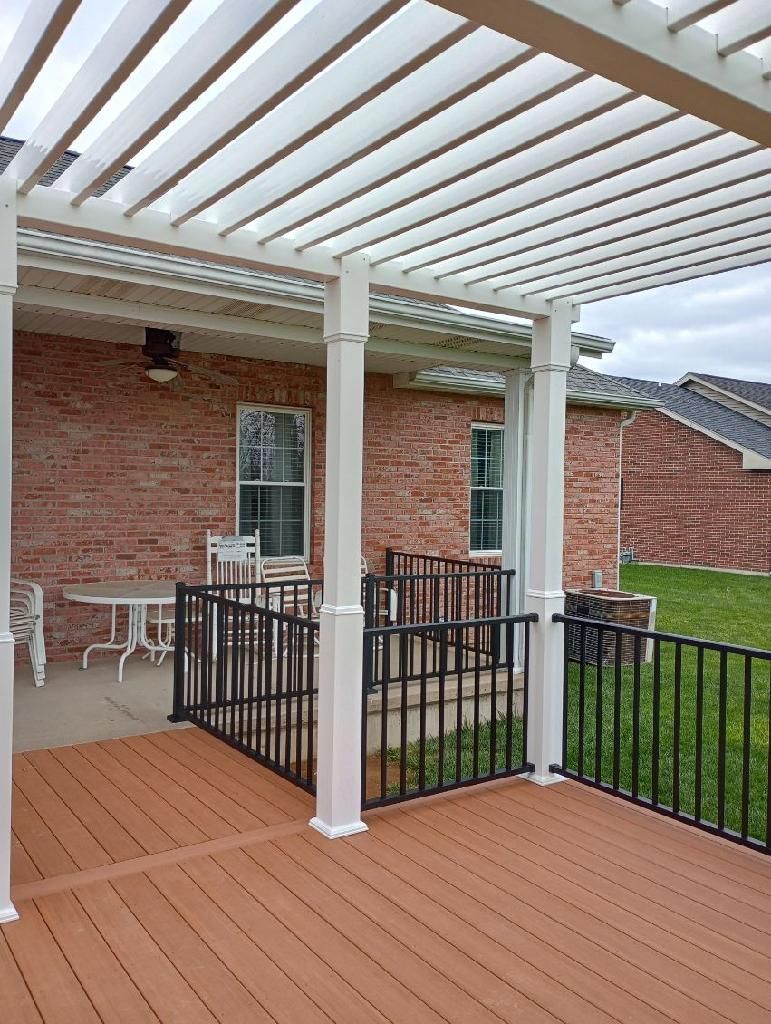 Covered wooden deck with white pergola, black railing, and brick house patio.