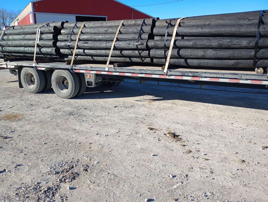 Flatbed trailer loaded with long black pipes, strapped down in a gravel lot near a red building.