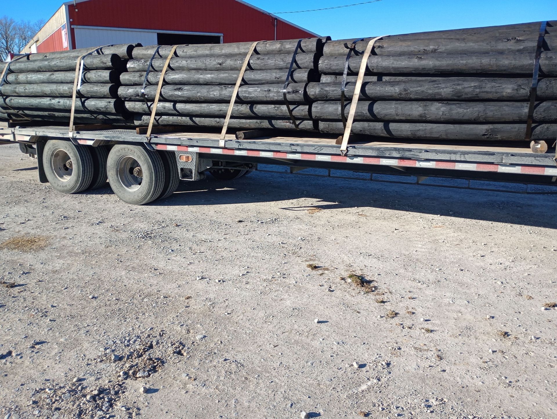 Flatbed trailer loaded with long stacked logs in a gravel yard, with a red building in the background