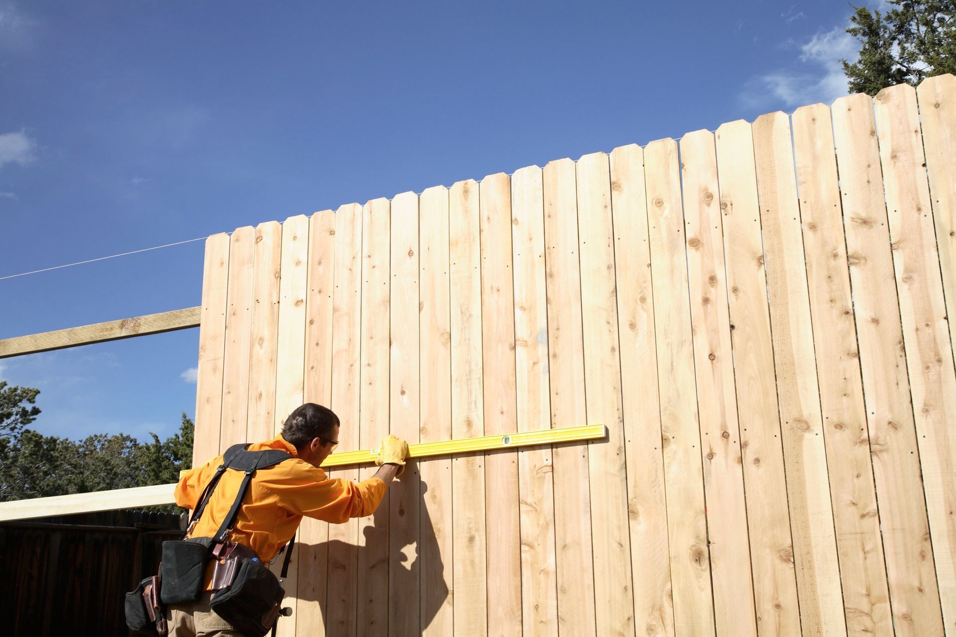 Worker measuring a tall wooden fence with a yellow tape measure under a blue sky
