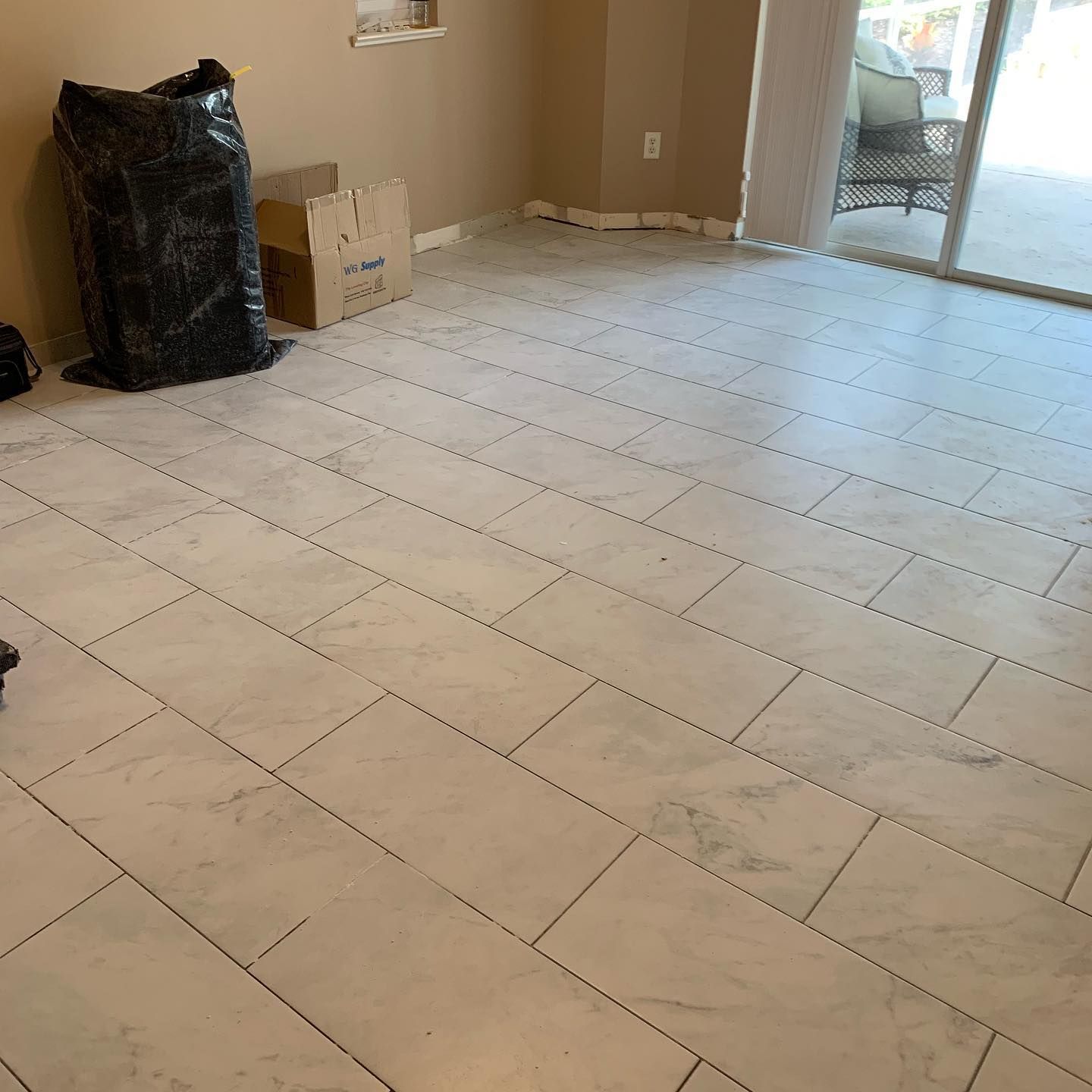 Empty tiled room with beige walls, a black bag in the corner, and an open doorway to the outside.