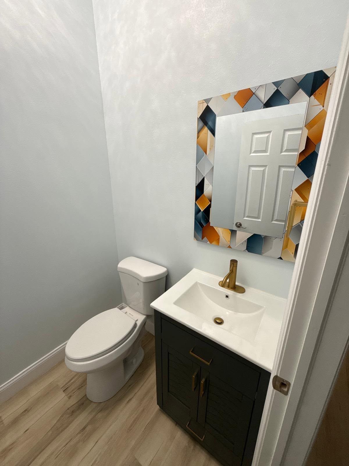 Small bathroom with toilet, sink vanity, and decorative mirror on light gray walls and wood-look floor.