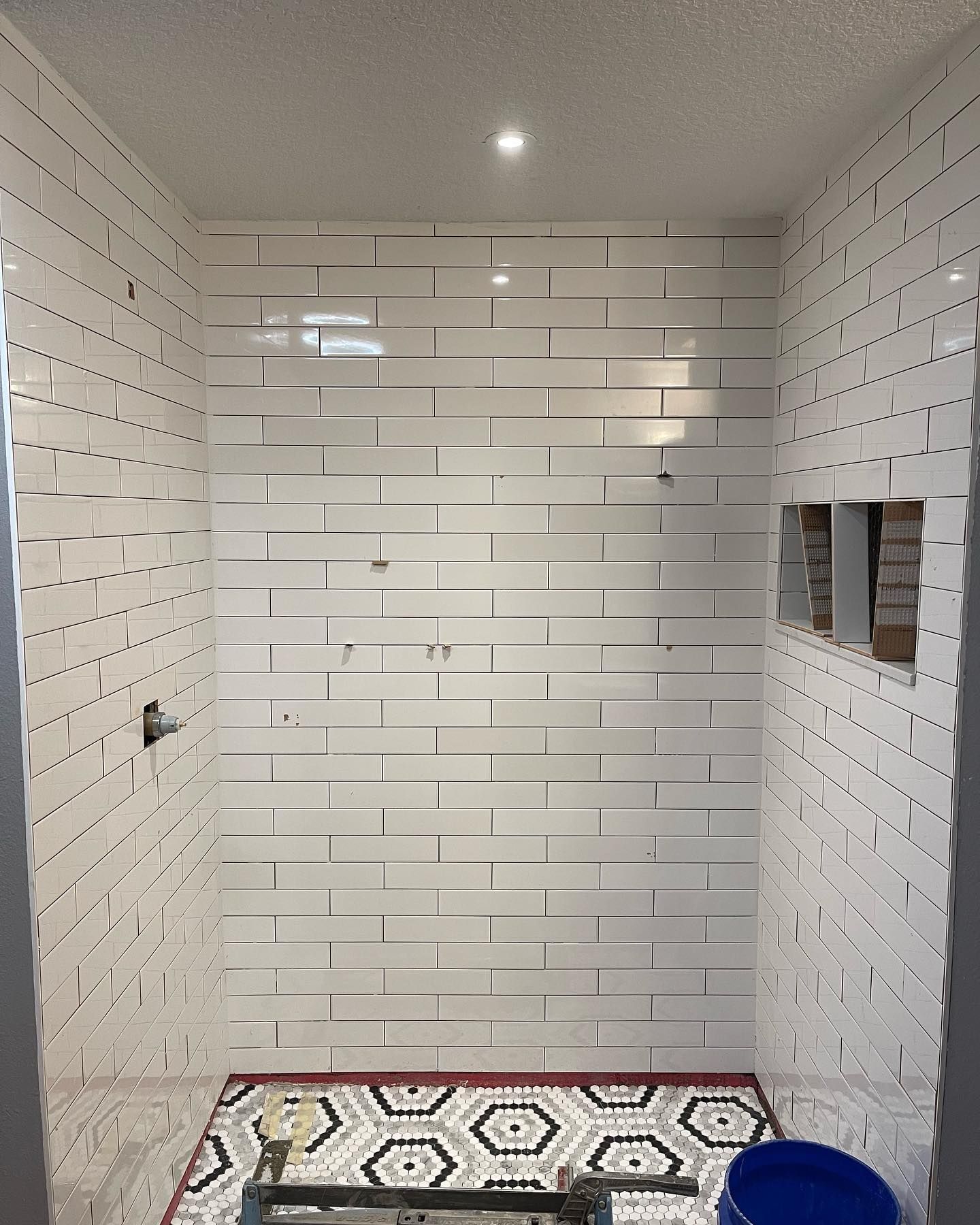 Small white-tiled shower stall with patterned floor mat and a blue bucket in the corner.