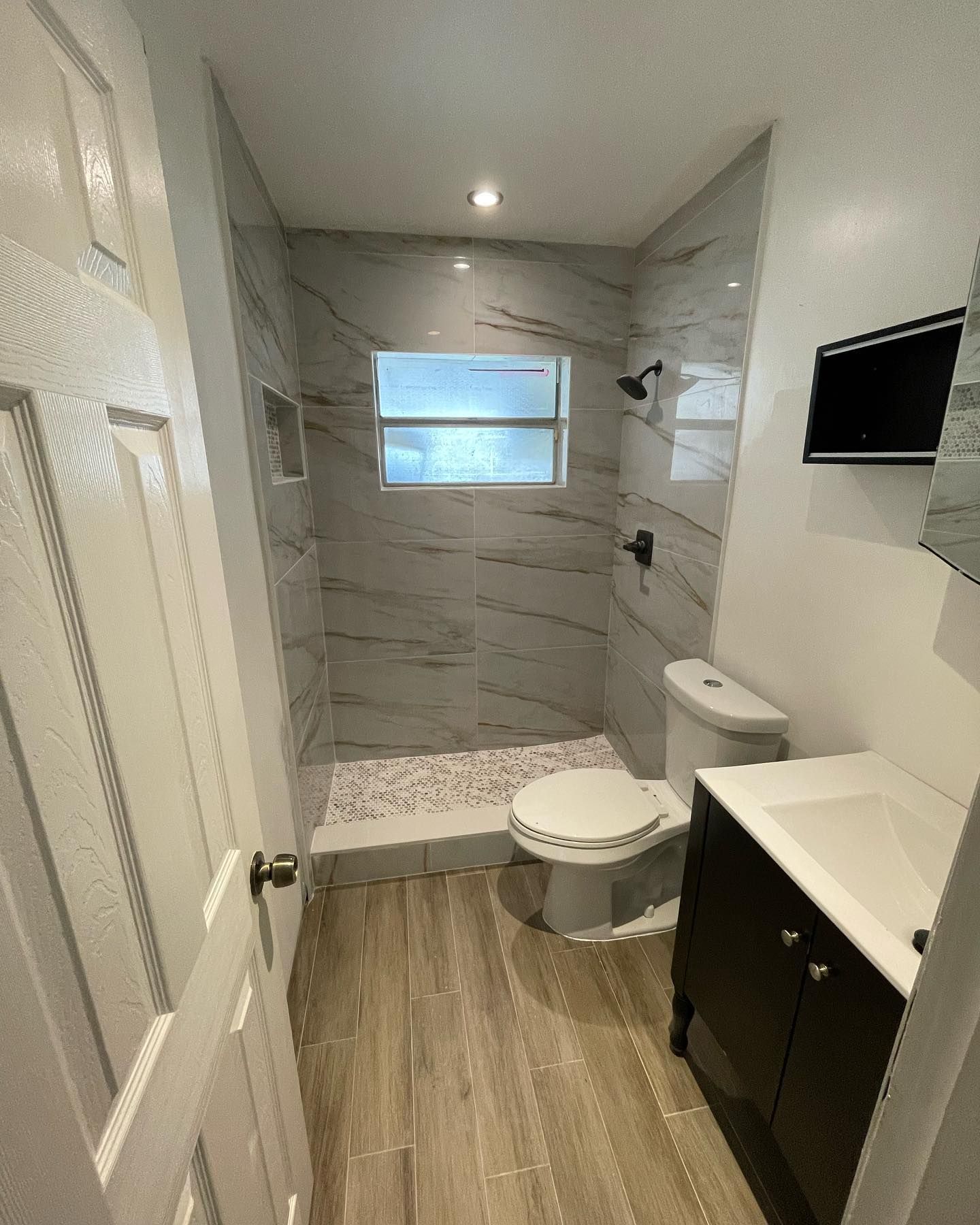 Compact modern bathroom with walk-in shower, toilet, vanity, wall-mounted TV, and wood-look floor