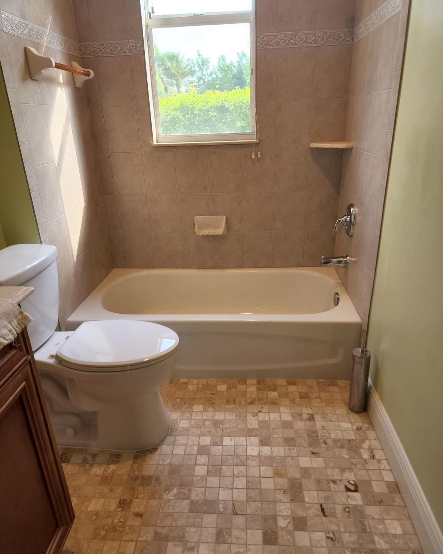 Small beige bathroom with tub, toilet, window, and mosaic tile floor.