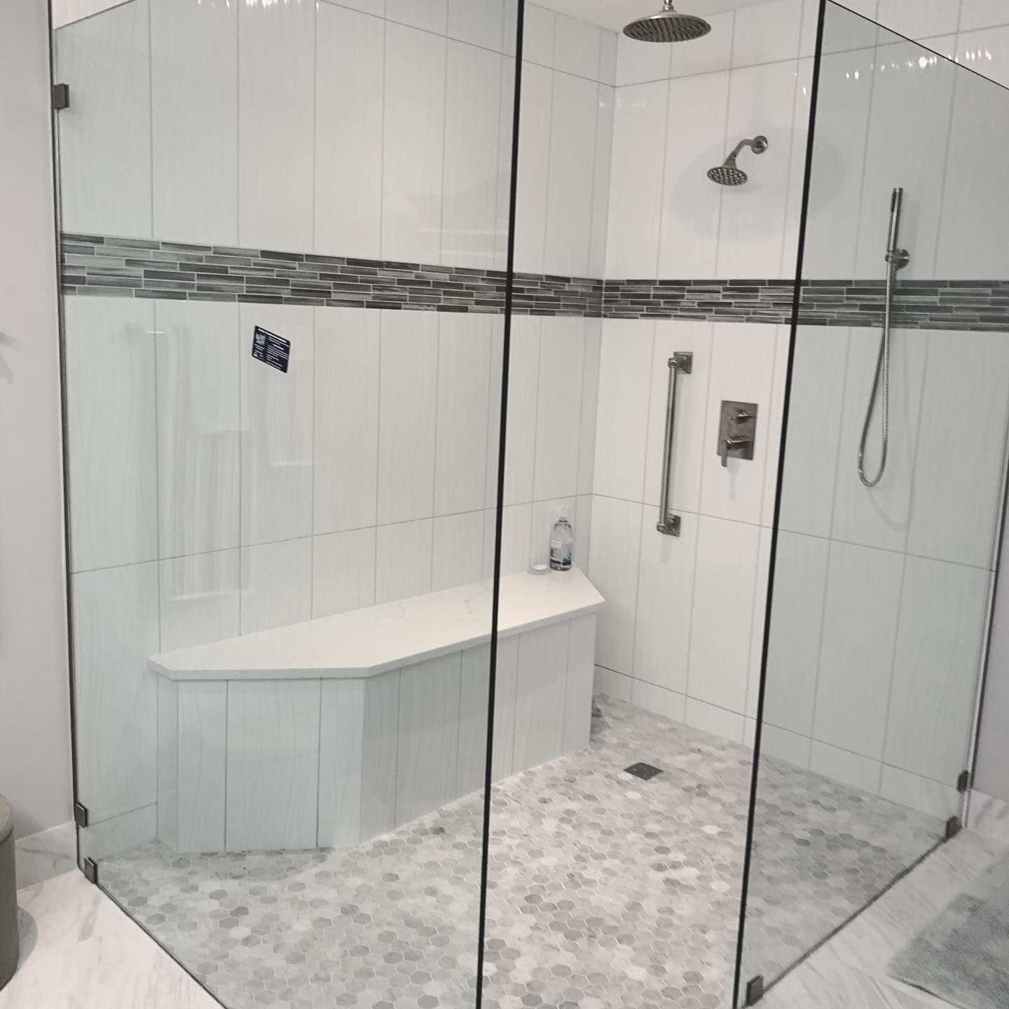 Accessible walk-in shower with glass enclosure, white tile walls, gray mosaic floor, bench, grab bar, and handheld showerhead