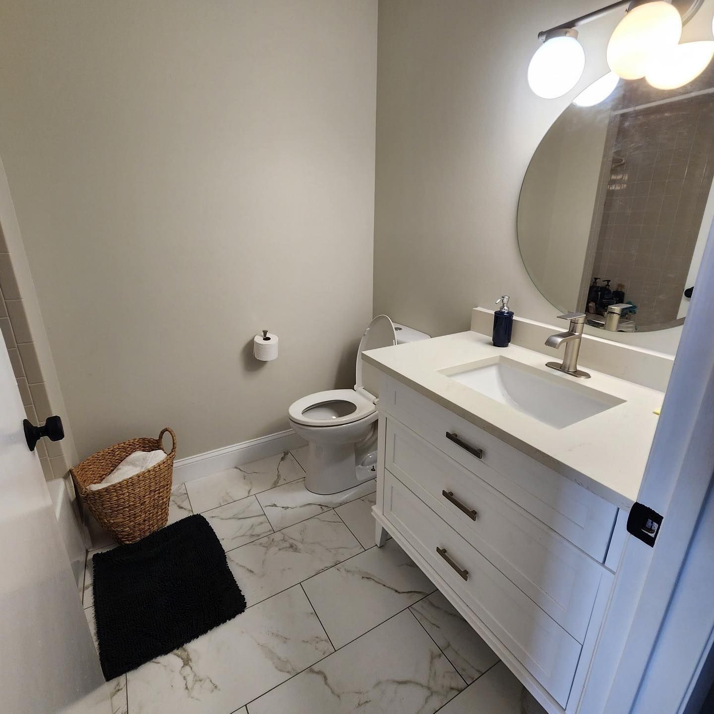 Small bathroom with vanity, mirror lights, toilet, woven basket, and black floor mat.