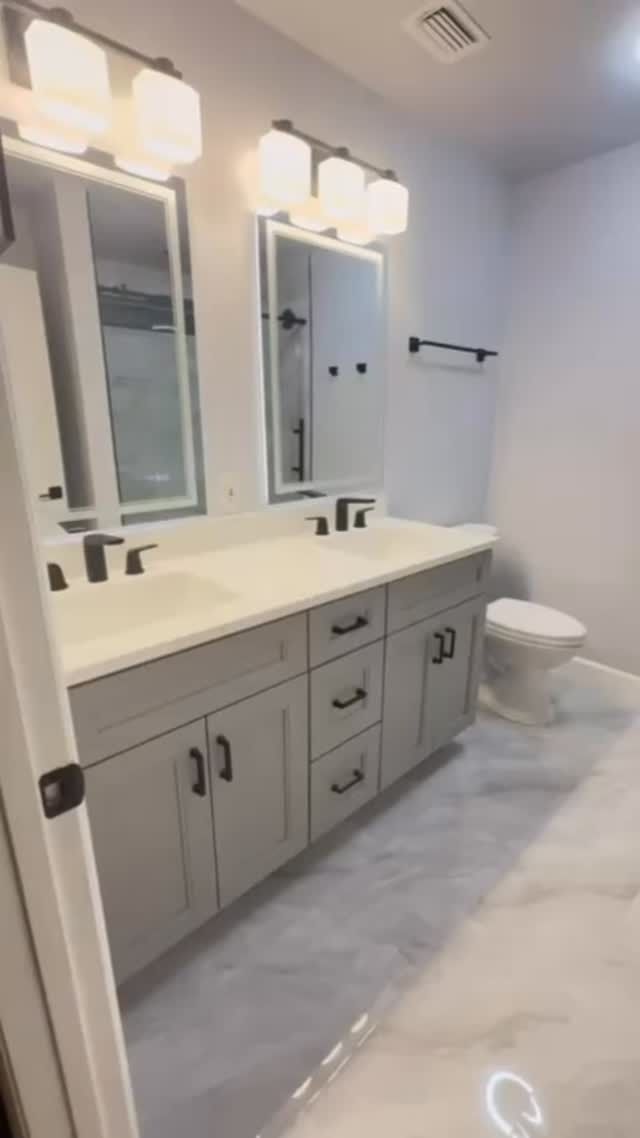 Bright modern bathroom with double vanity, large mirrors, white cabinets, marble floor, and toilet.