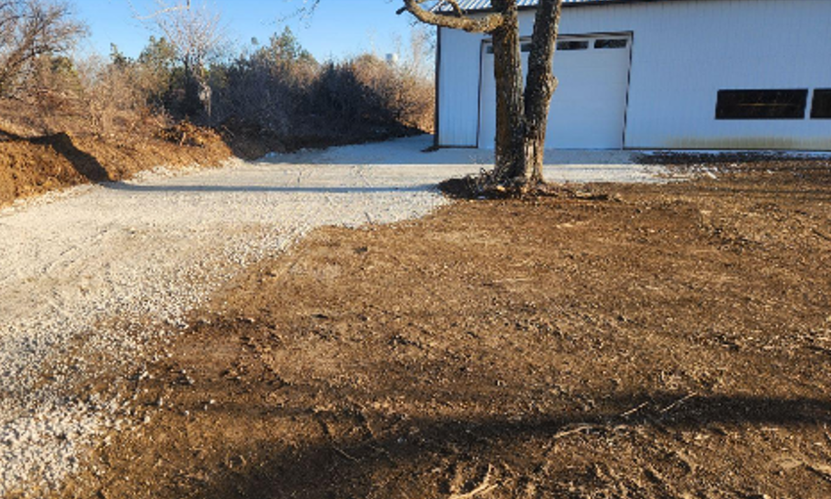 Gravel driveway leading to a white building. A tree is to the right with dirt and grass in the foreground.
