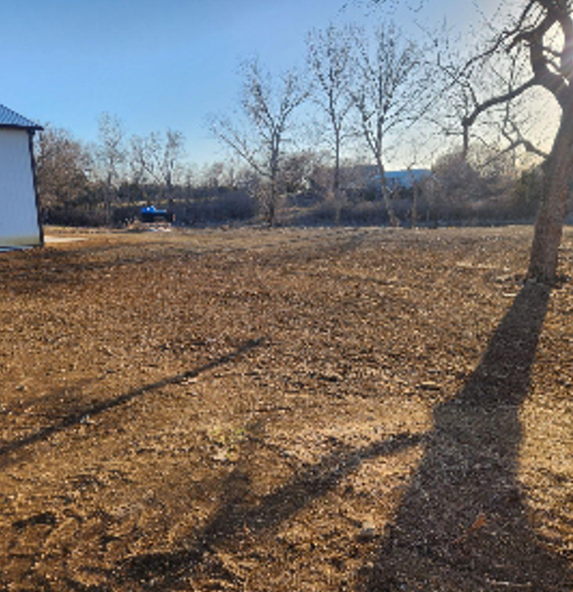 An empty, brown field under a blue sky, with bare trees in the background. A building is visible on the left.