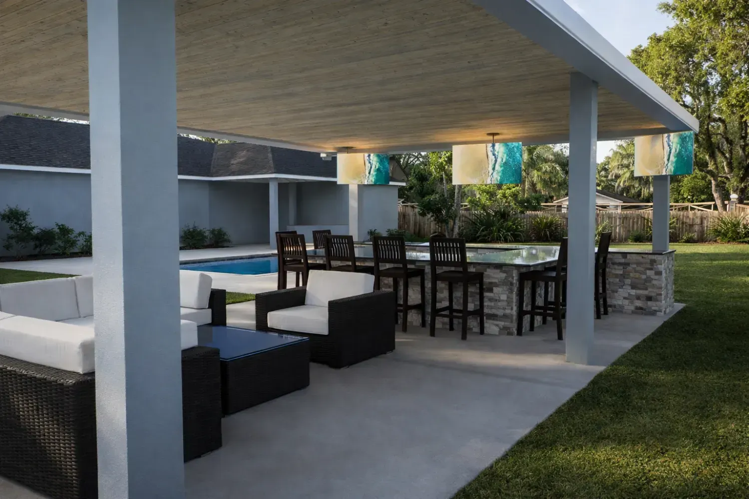 Outdoor patio with a covered seating area, pool, and a stone bar with stools, all set on a concrete slab near a lawn.