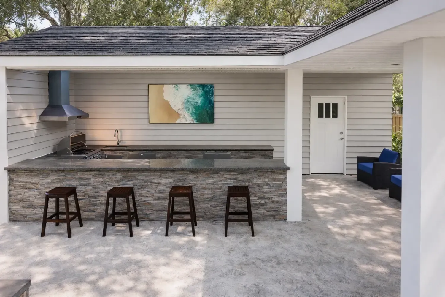 Outdoor kitchen with a stone bar, four dark stools, a vent hood, and wall art under a covered patio.