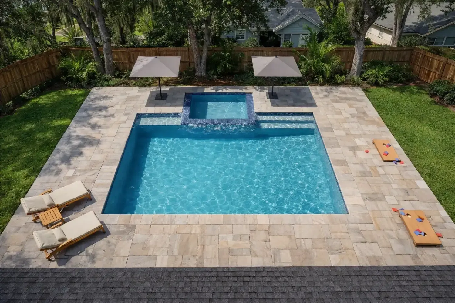 A square swimming pool with an integrated spa, surrounded by stone pavers, lounge chairs, and two wooden lawn games.