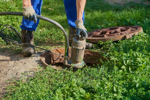 Worker operating a submersible pump and hose at an access pit in a grassy outdoor area. Worker operating a submersible pump and hose at an access pit in a grassy outdoor area.