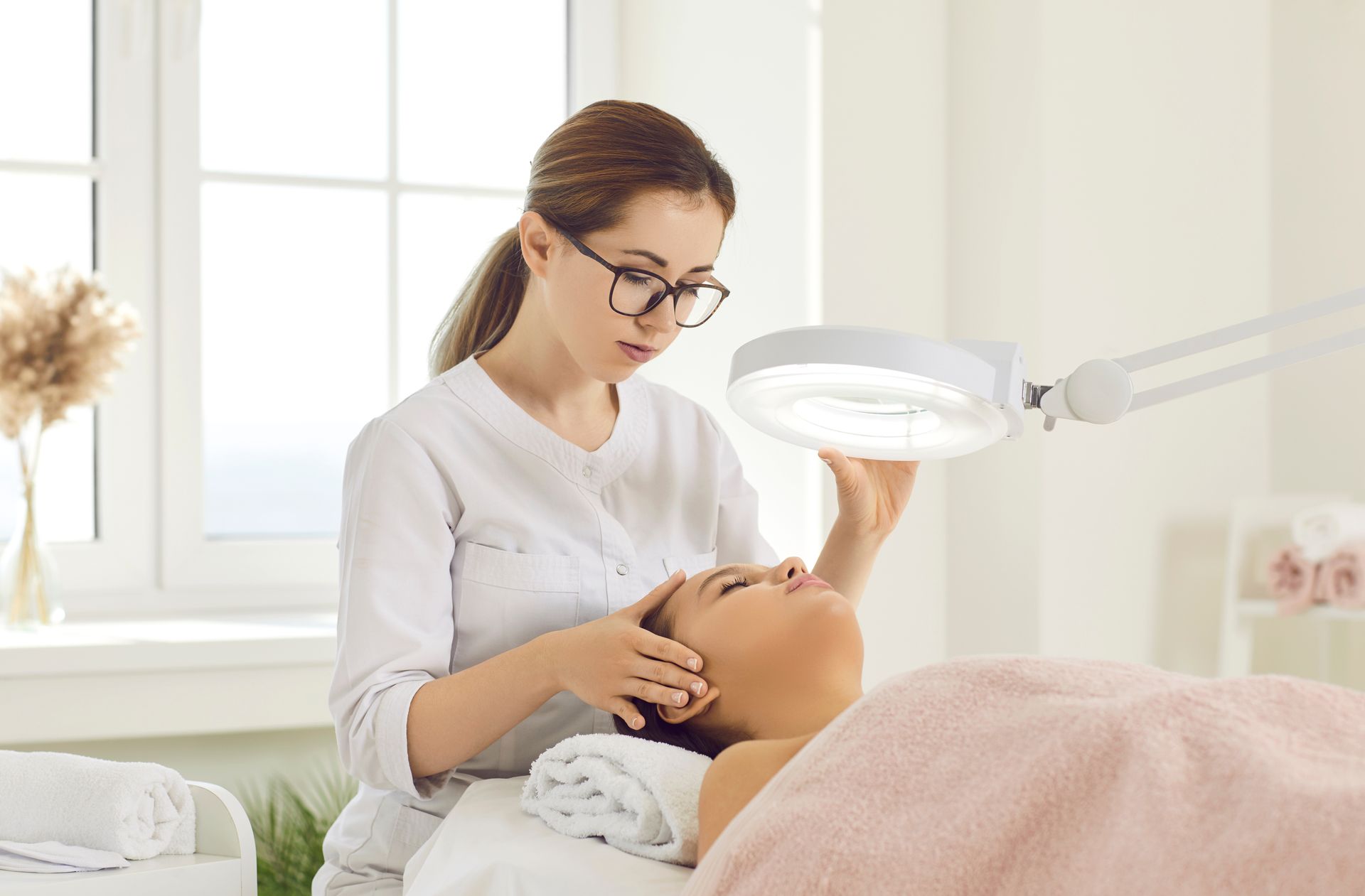 A woman closely examines a patient's skin during dermatology treatment under a lighted magnifier.