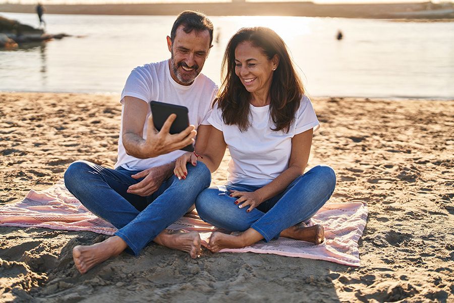 Couple sits cross-legged on a beach, smiling at a tablet. Sunlight. Sandy beach.