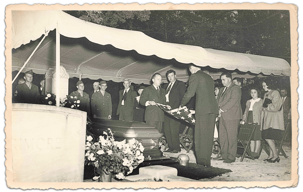 People at a funeral, placing a wreath on a casket under a canopy. Black and white photo.