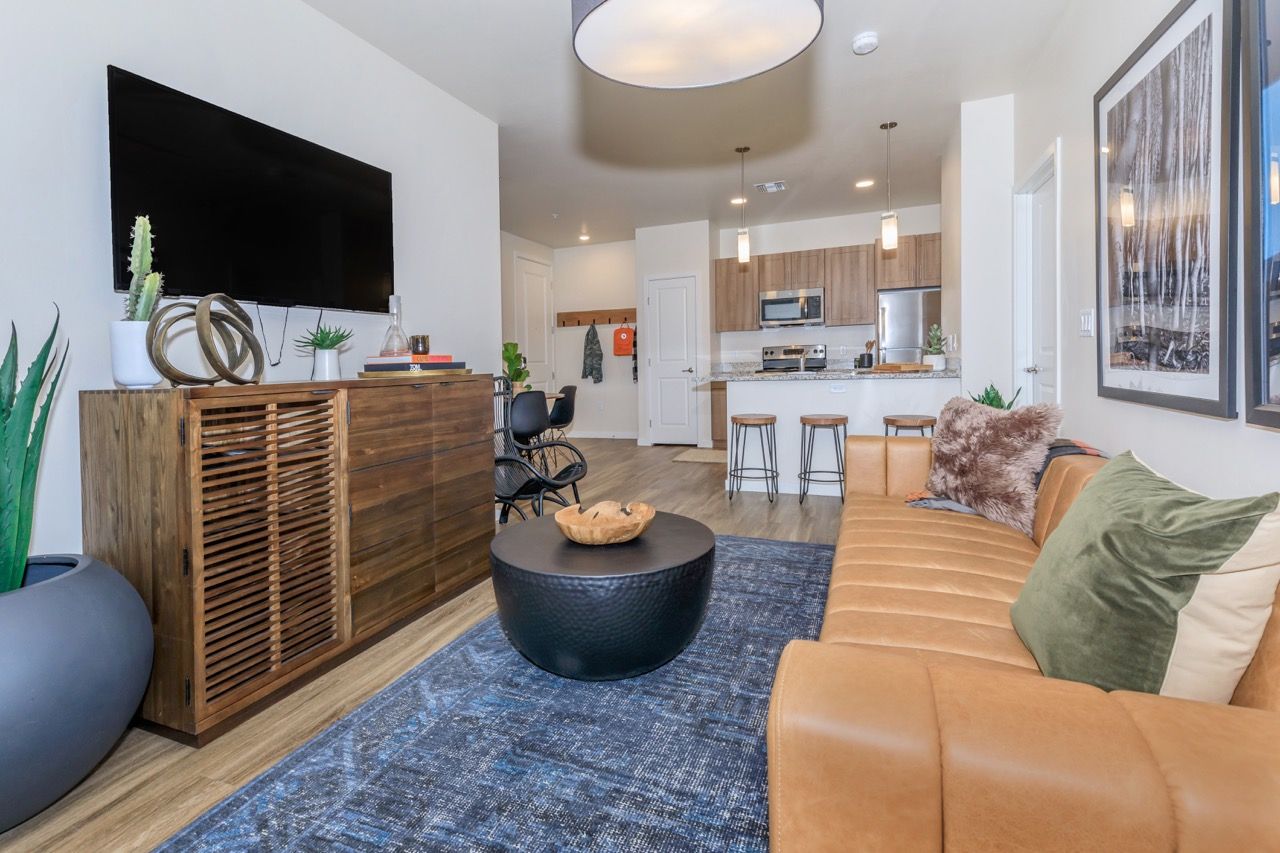 Open living area with a tan sectional sofa, coffee table, TV, and kitchen in the background.