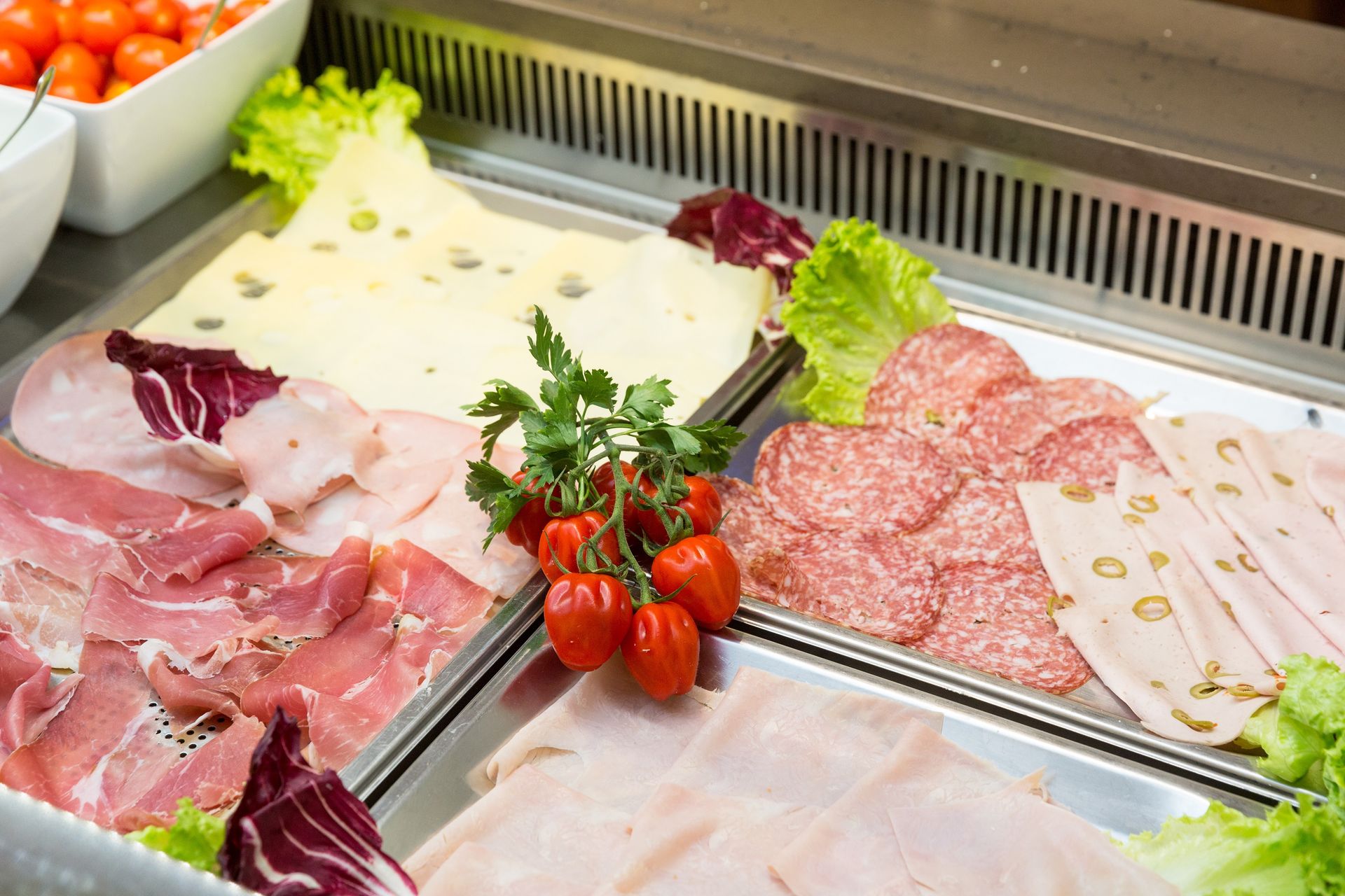 Assorted deli meats and sliced charcuterie on a stainless-steel buffet counter with tomatoes and lettuce garnish