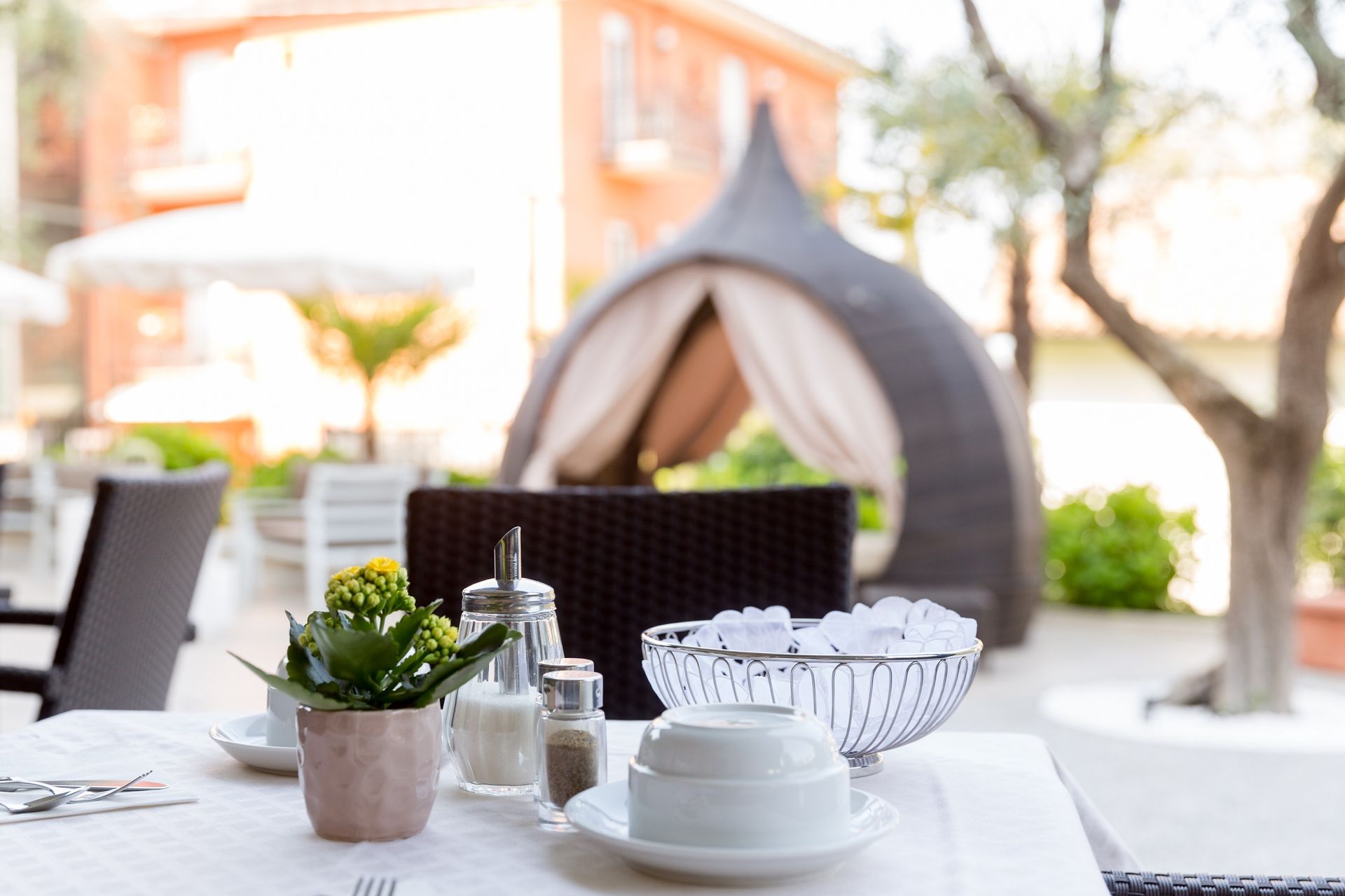 Outdoor patio table set for dining, with plants, dishes, and a tent-like lounge in the background