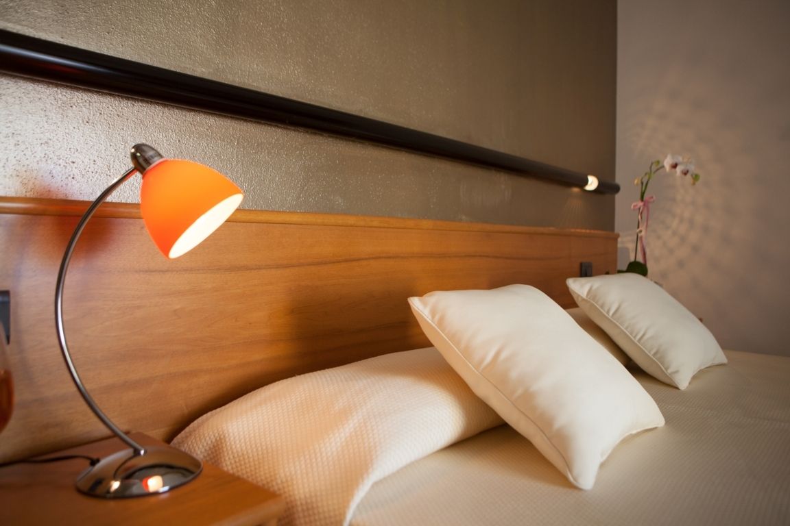 Letto d'albergo con cuscini bianchi, testiera in legno e lampada da comodino arancione a sinistra.