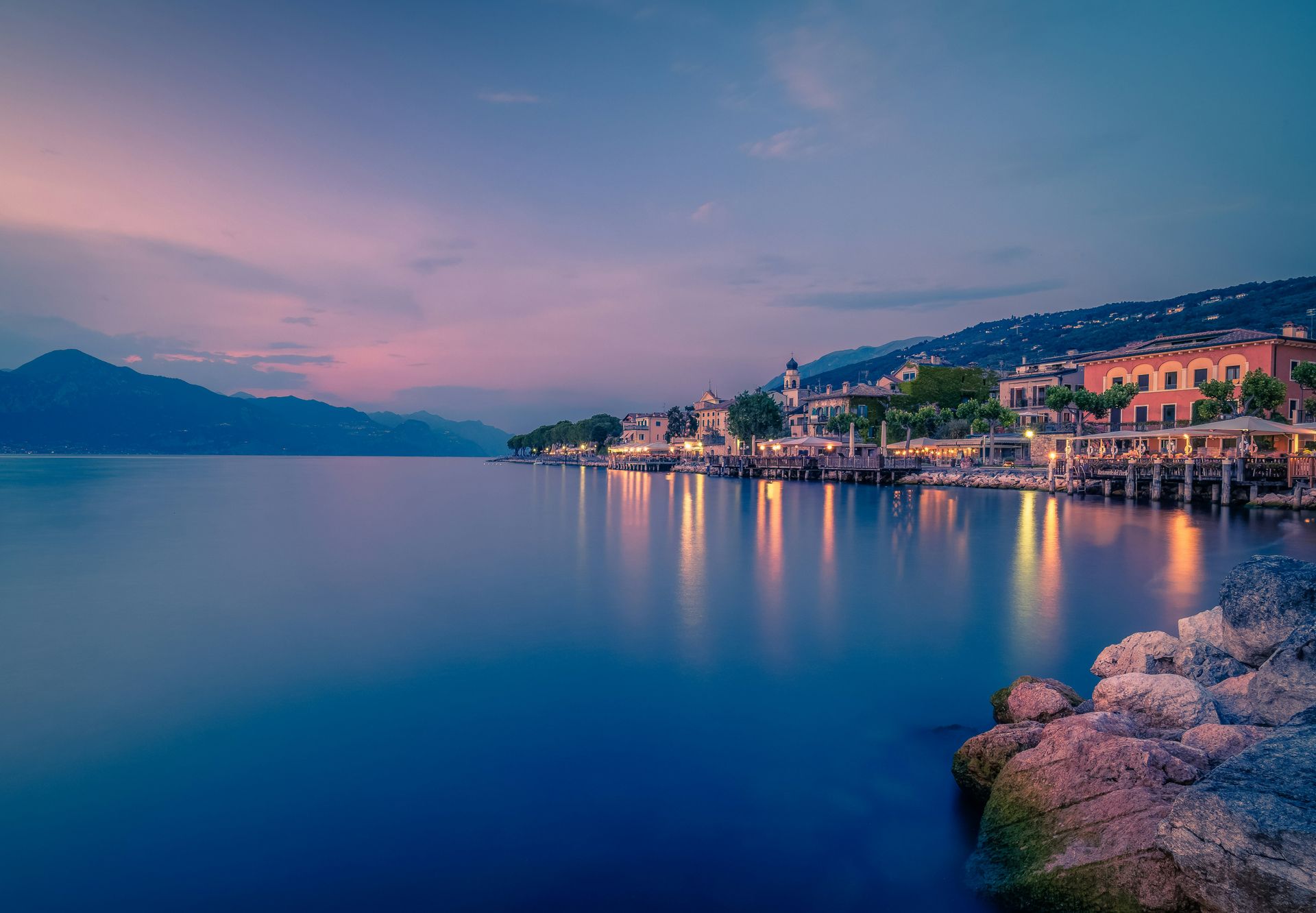 Calm lakeside town at dusk with glowing waterfront lights and mountains across a still bay