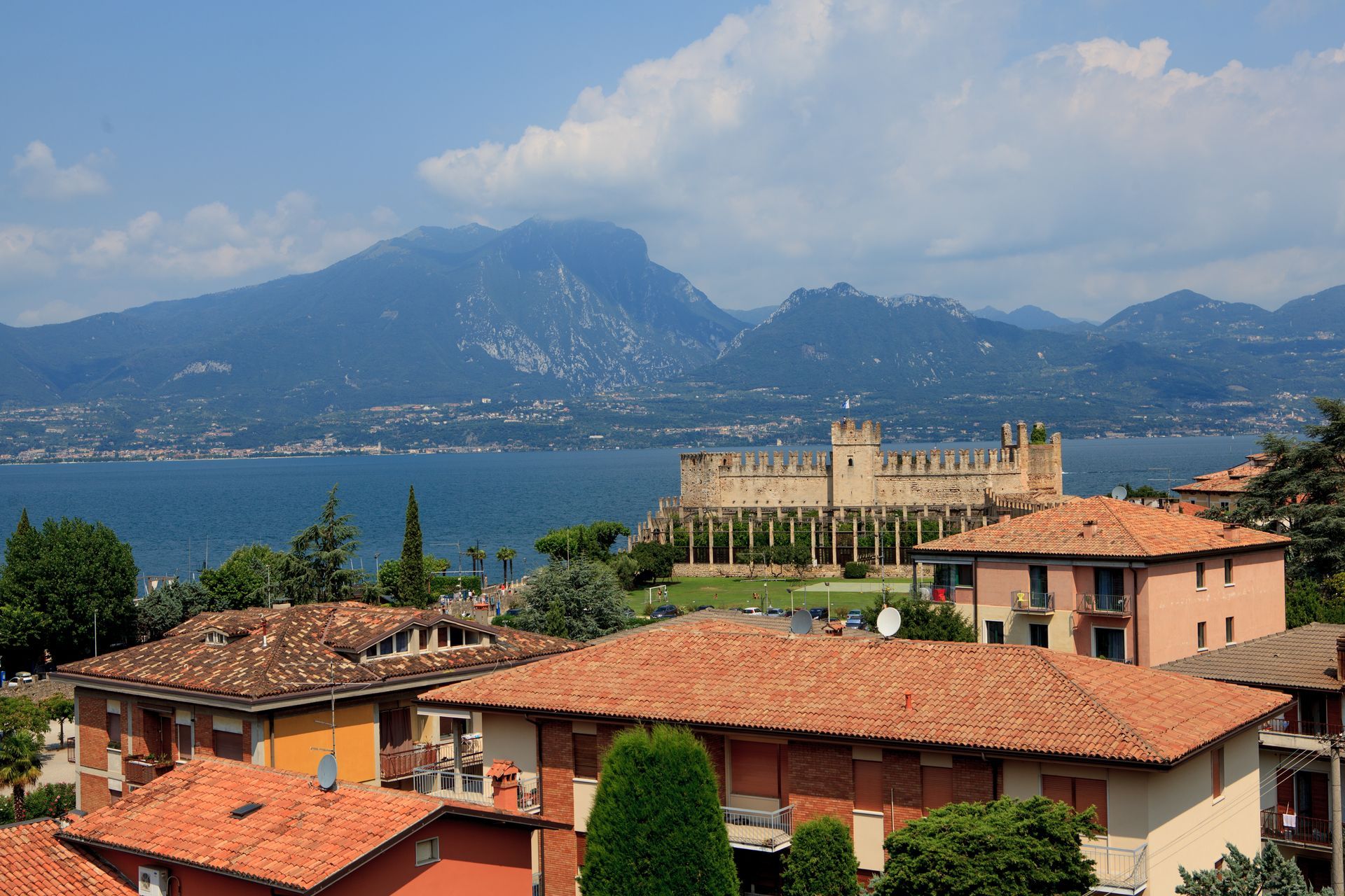 I tetti di un villaggio sul lago con un castello e le montagne sullo sfondo, sotto un cielo nuvoloso.