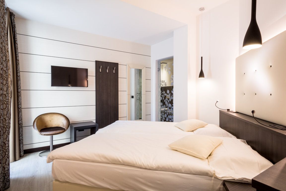 Modern hotel room with a bed, wall-mounted TV, desk, chair, and black pendant lights