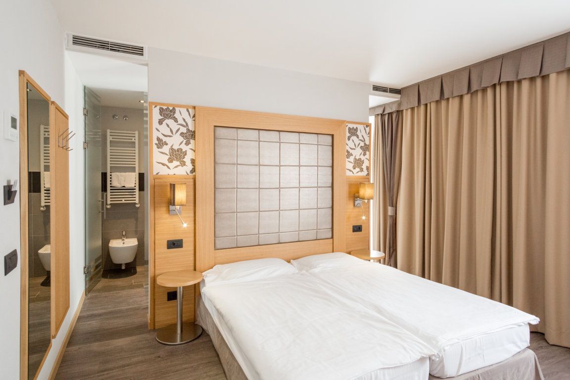 Modern hotel bedroom with white bed, wooden headboard, beige curtains, and en suite bathroom glimpse