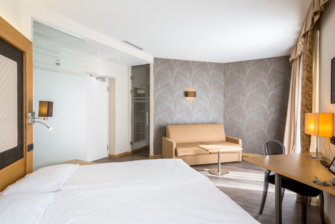 Bright hotel room with bed, beige sofa, desk, and patterned accent wall near a window