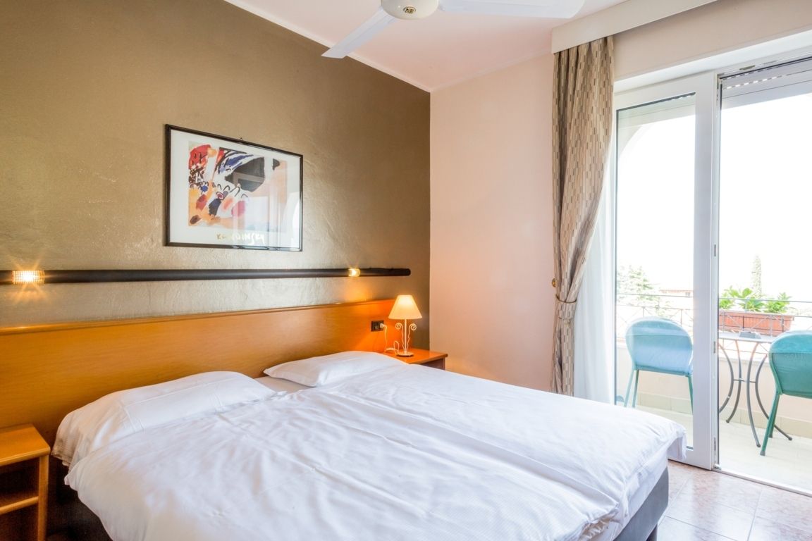 Bright hotel room with a double bed, orange headboard, and balcony doors to a small table and chairs.