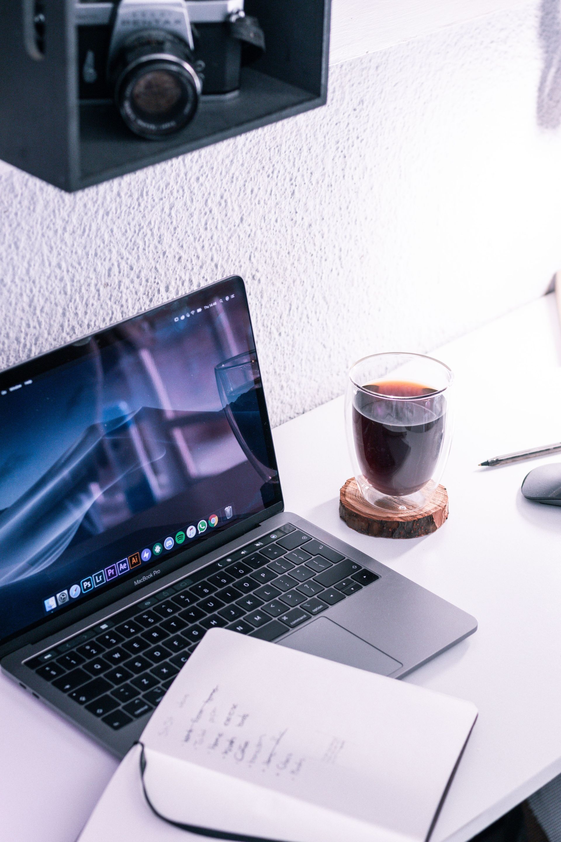 Laptop on white desk with notebook and iced coffee beside it, under a wall shelf.