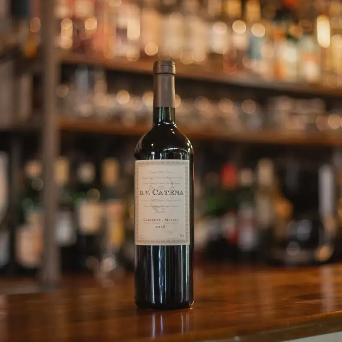 Una botella de vino tinto DV Catena junto a una copa sobre una mesa iluminada con luz cálida.