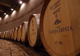 Filas de grandes barricas de madera para vino en una bodega, etiquetadas como Catena Zapata bajo una iluminación cálida.