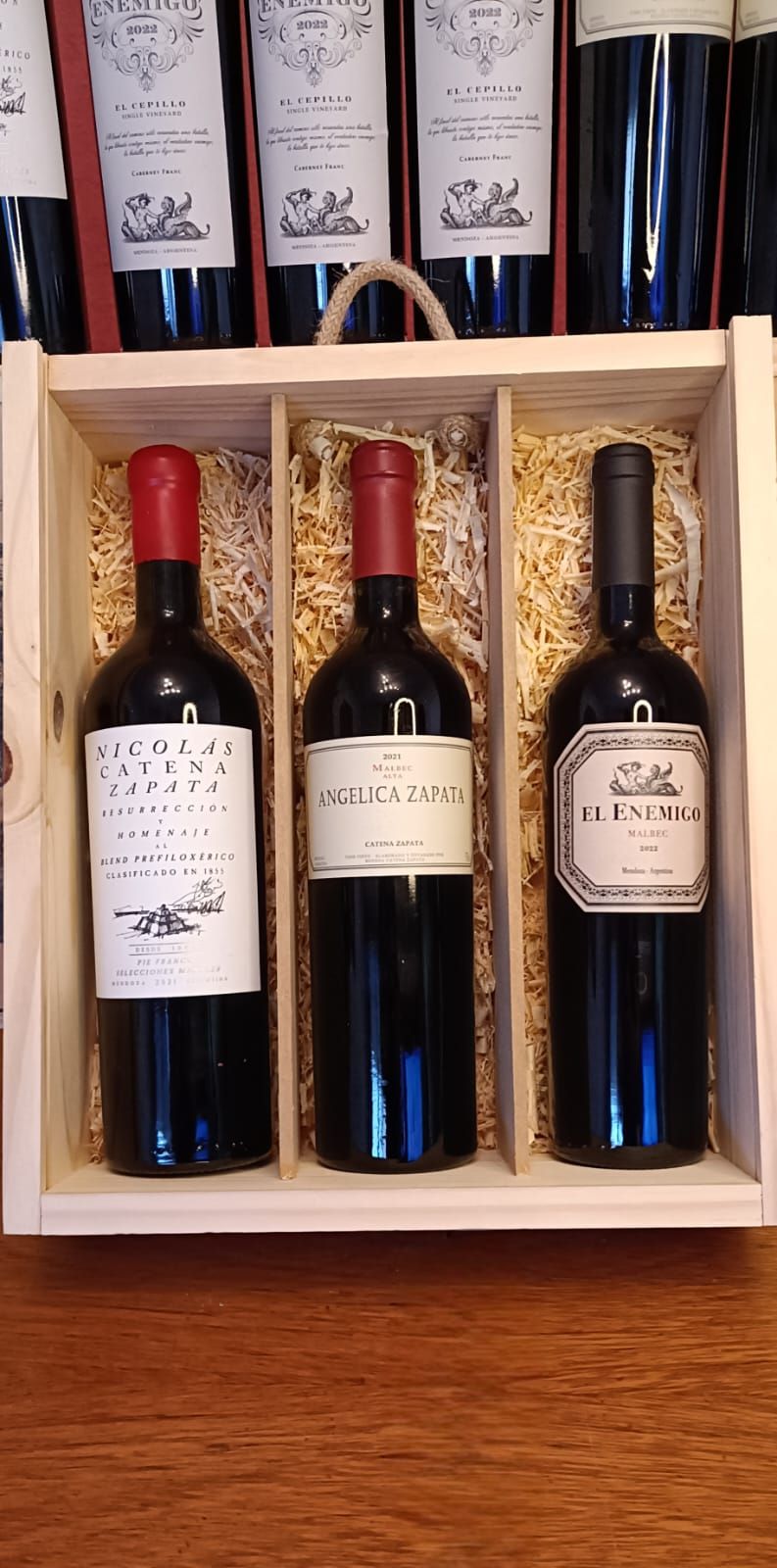 Tres botellas de vino tinto en una caja de regalo de madera con botellas azules al fondo.