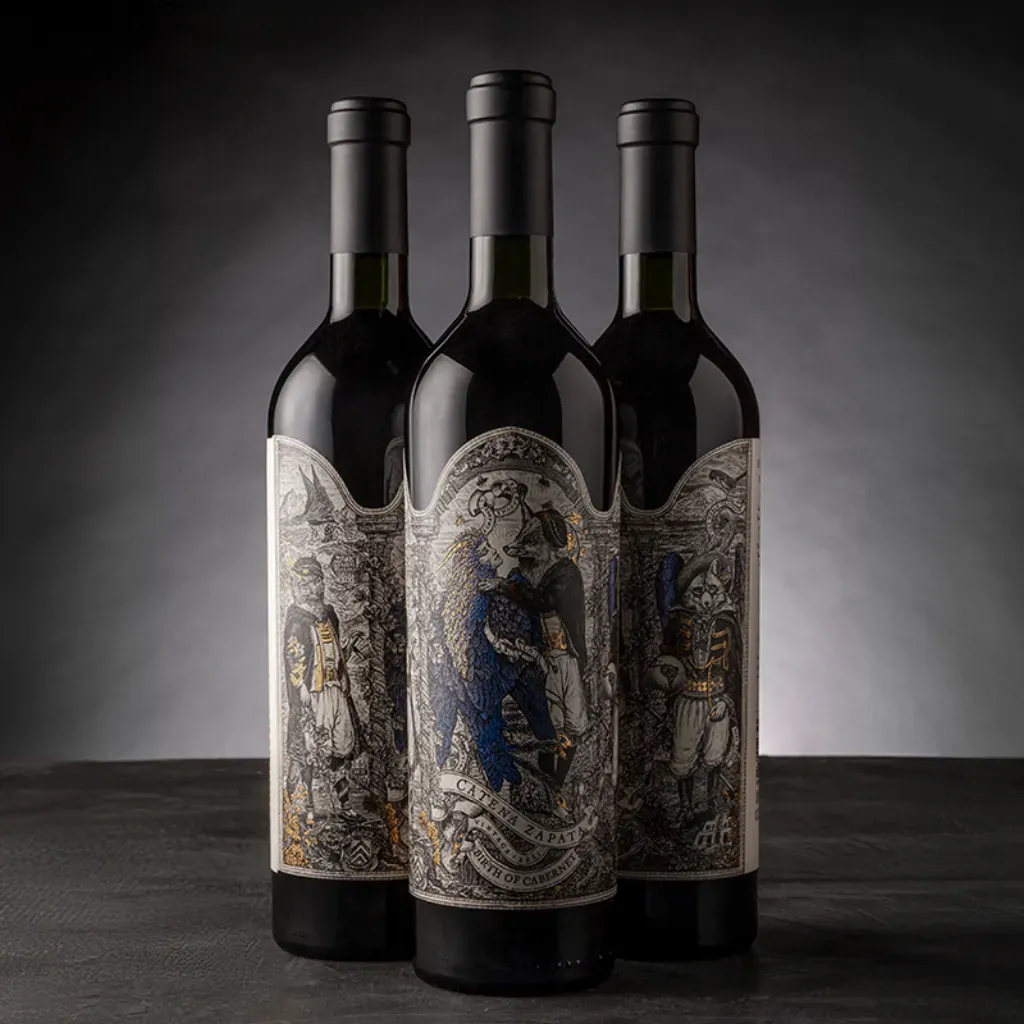Tres botellas de vino oscuro con etiquetas ornamentadas dispuestas sobre una mesa negra sobre un fondo gris.