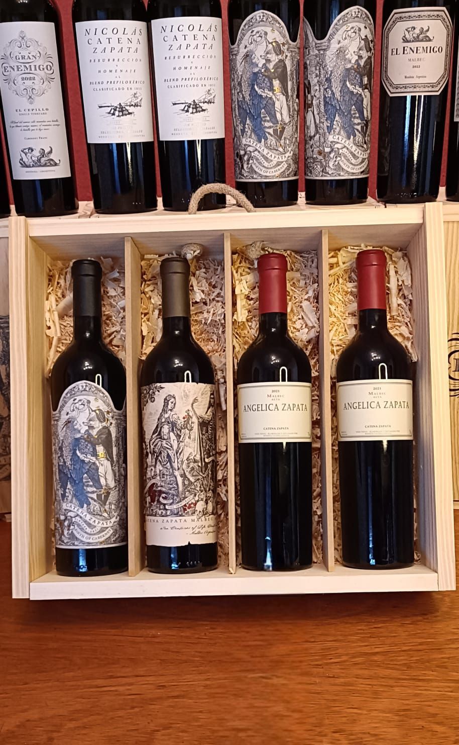 Caja de madera con botellas de vino tinto surtidas sobre una mesa, algunas con papel de aluminio azul y etiquetas blancas.