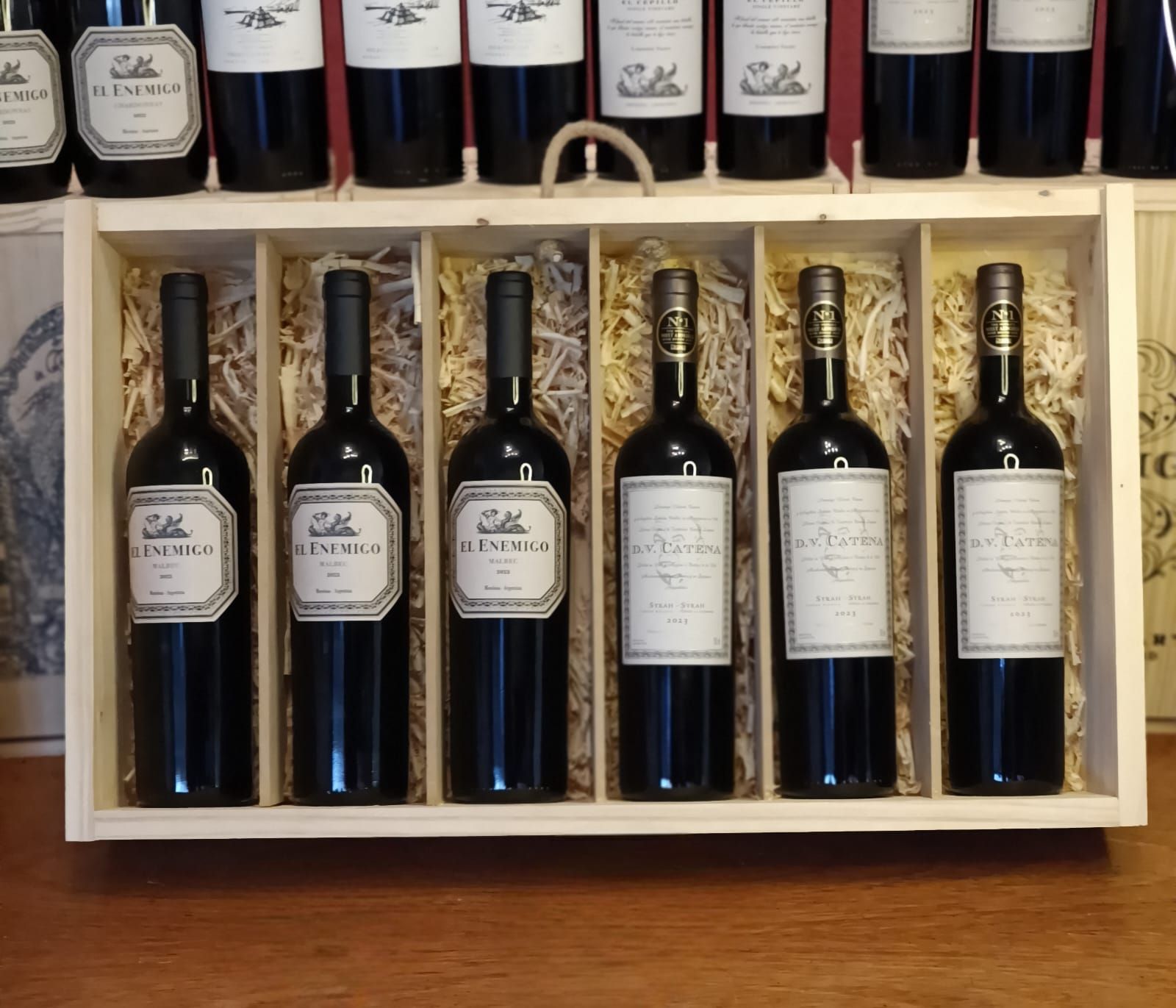 Expositor de madera para vinos con seis botellas en una caja y más botellas en un estante superior.
