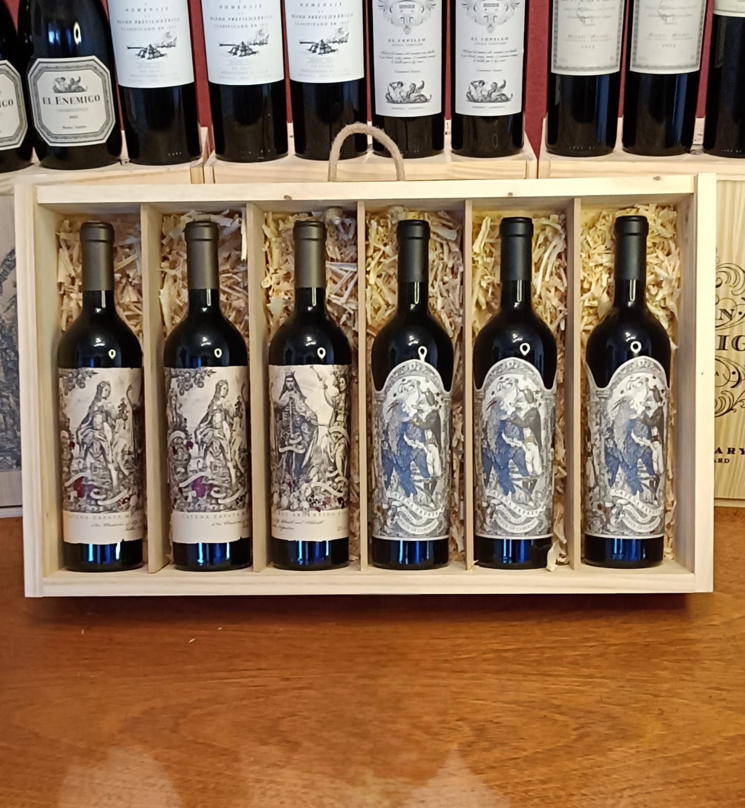 Una caja de madera con cinco botellas de vino oscuro sobre una mesa, con más botellas expuestas en un estante detrás.